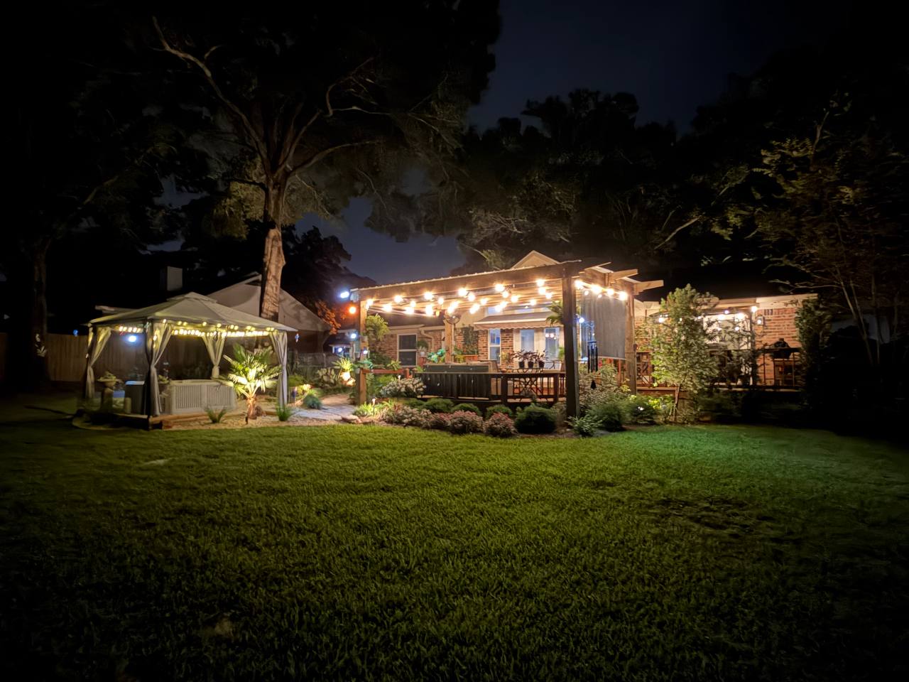Back garden at night with a lit pergola, spa gazebo, layered planting beds, and broad lawn in the foreground