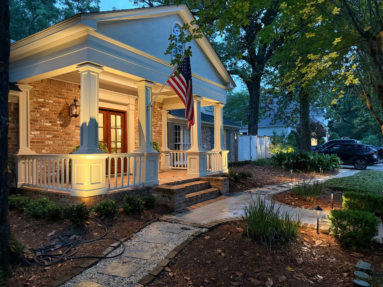 Angled dusk view of a brick home with white-columned porch, path lighting, and layered planting along the side walk