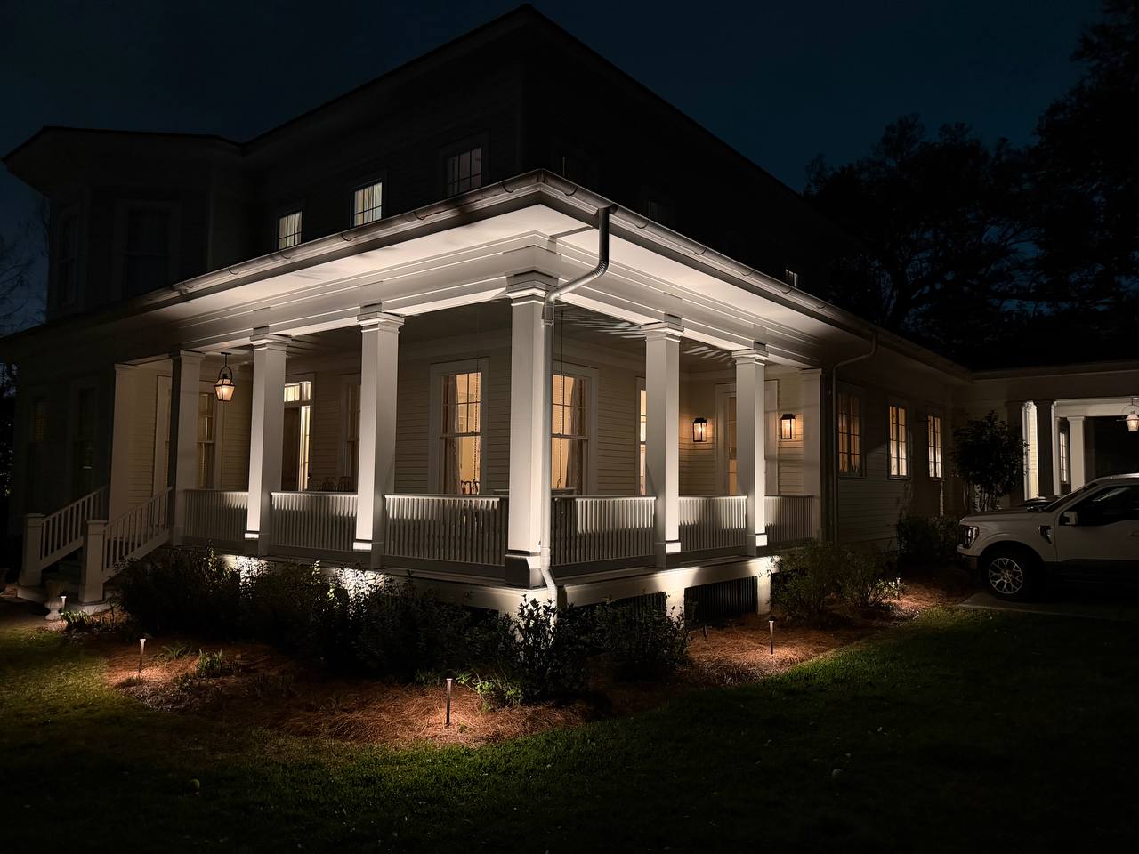 Wraparound porch at night with crisp white columns, warm underlighting, and soft foundation planting below