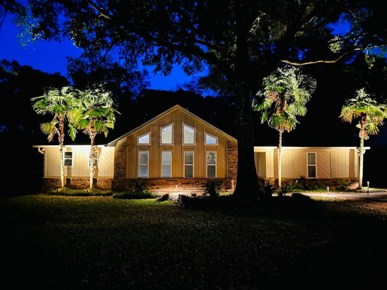 Low modern home with tall palms, a large oak, and warm facade lighting across the front lawn at blue hour